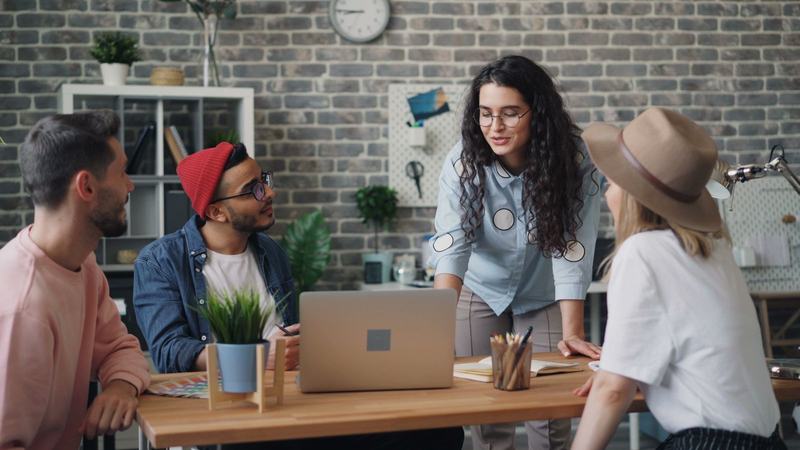 A small group gathered around a table in a bright office, talking while one woman stands and leads the conversation