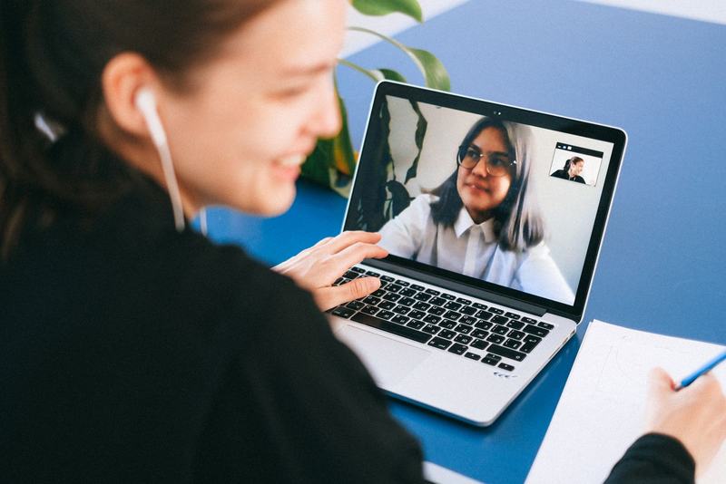A smiling woman wearing earphones talks with someone on a laptop video call while taking notes